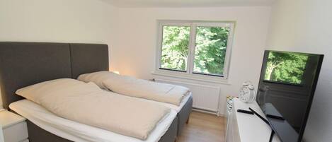Une chambre à coucher comprenant un lit, des tables de chevet, un bureau avec un ordinateur et une fenêtre donnant sur des arbres.