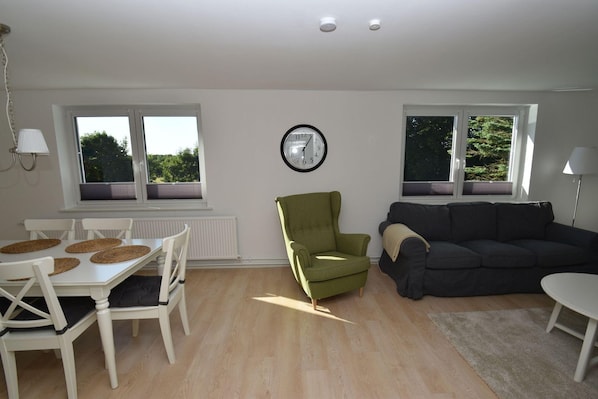 A living room with a dining table, a green armchair, a dark sofa, and a clock on the wall. - fewo1846 - Solskin - fewo1846 - Solskin / comfortable 3-room apartment in a rural location (Flensburg)