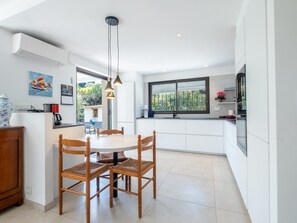 Una cocina moderna con muebles blancos, una mesa de comedor con cuatro sillas, iluminación colgante y una ventana con vistas a zonas verdes.