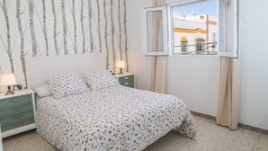 A bedroom with a bed, a nightstand, a lamp, a window with a view of buildings, and a wall with a tree pattern.