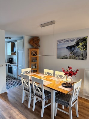 Ein Essbereich mit Holztisch und weißen Stühlen, im Hintergrund eine Küche und ein Küstenlandschaftsbild an der Wand.