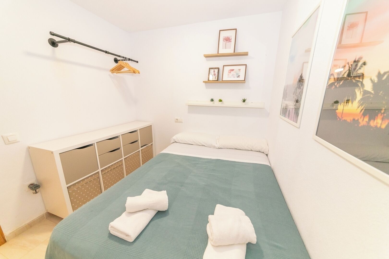 Un dormitorio con cama, armario, estante con fotografías y ventana con vista al atardecer.