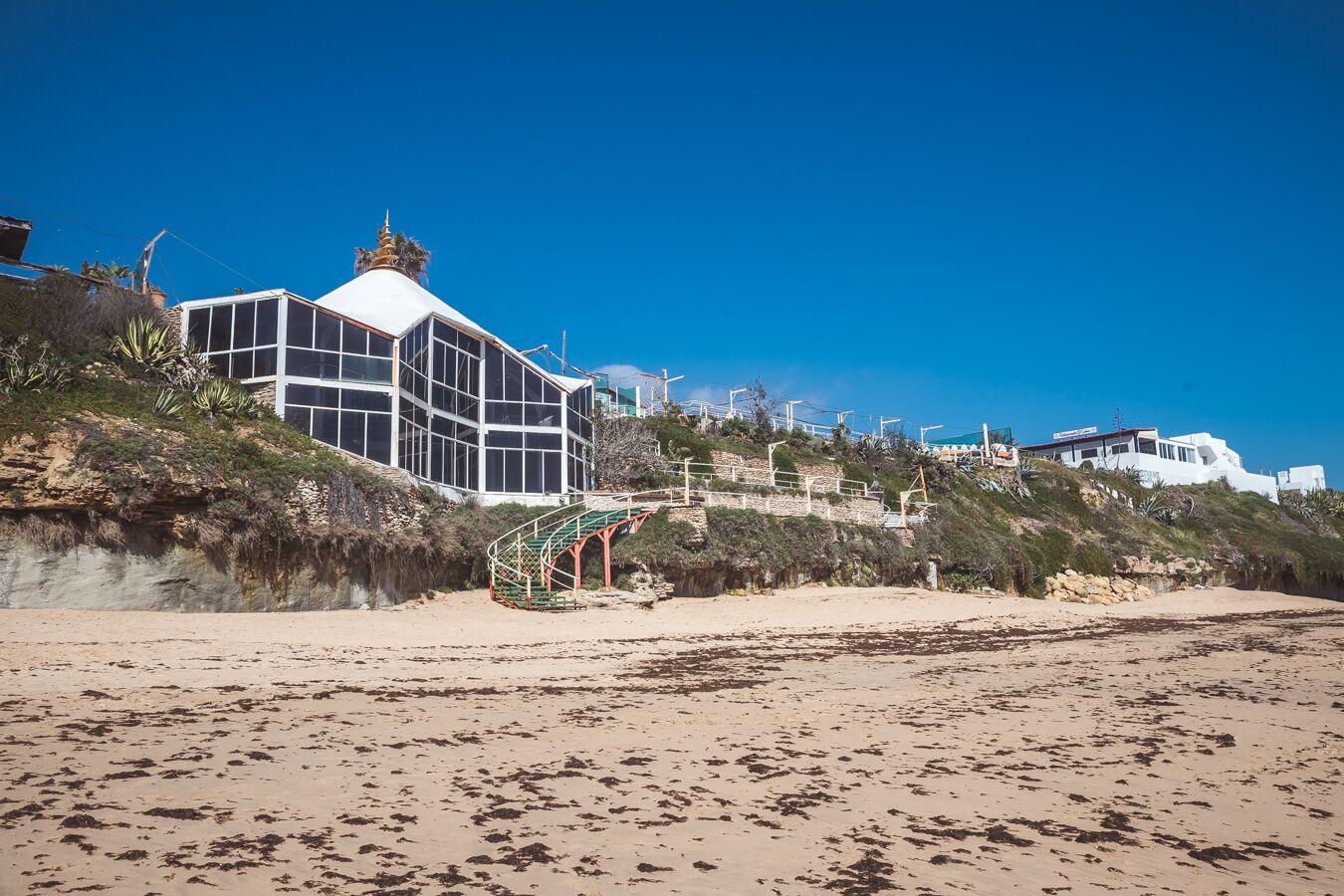A coastal building with a large glass facade, situated on a sandy beach with a staircase leading up to it.