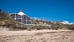 Ein Küstenhaus mit großer Glasfassade, gelegen an einem Sandstrand, mit einer Treppe, die zu ihm hinaufführt.