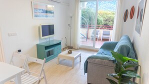 Ein modernes Wohnzimmer mit einer Couch, einem Fernseher, einem Esstisch und einem Balkon mit Blick auf Bäume und einen Berg.