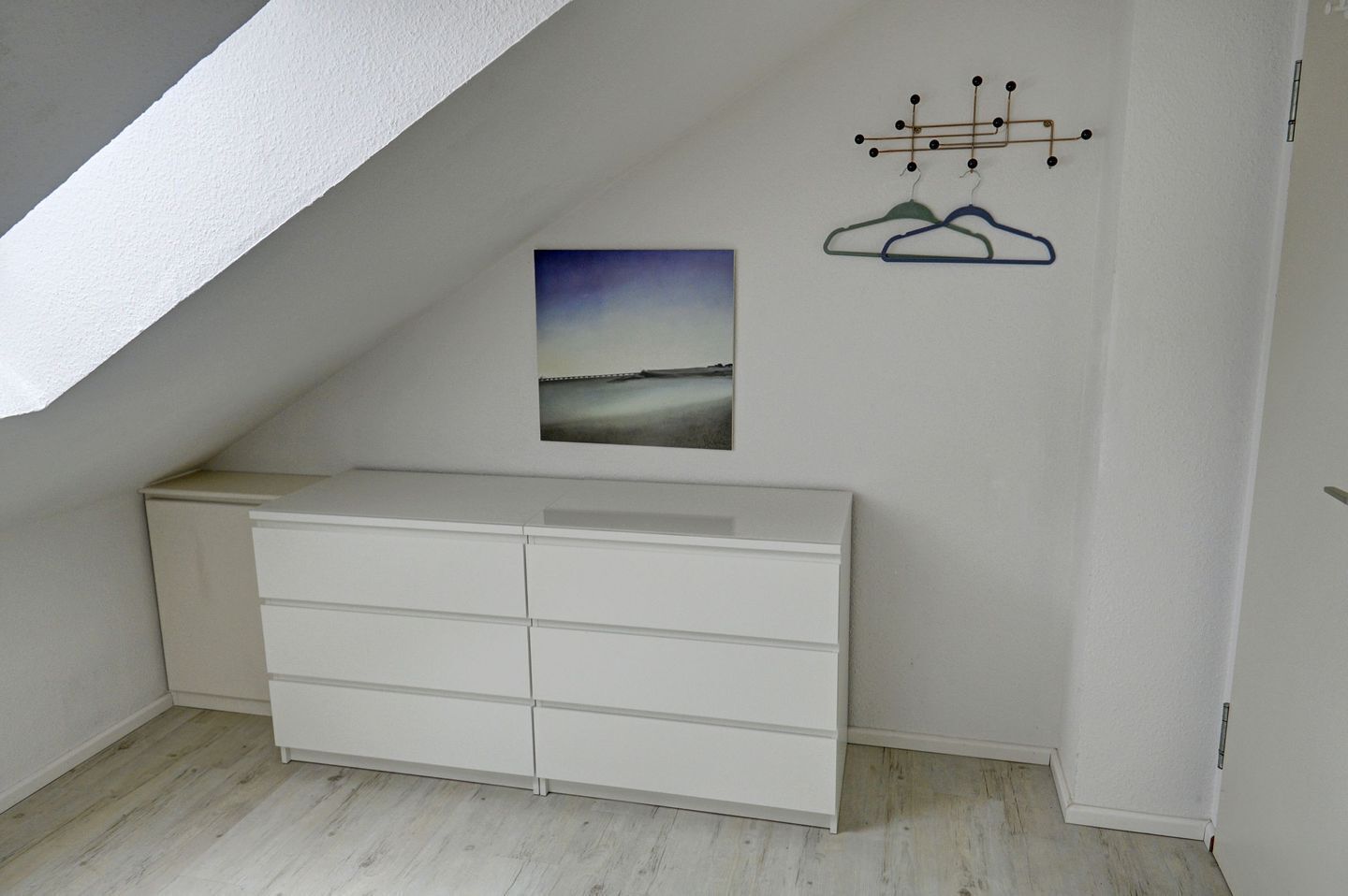 Une pièce avec une commode blanche, un tableau représentant un paysage et un porte-manteau avec des vêtements.