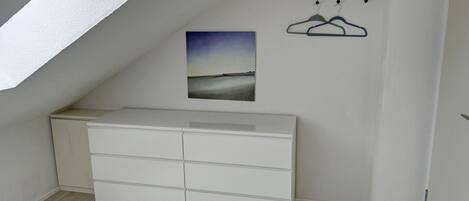 Une pièce avec une commode blanche, un tableau représentant un paysage et un porte-manteau avec des vêtements.