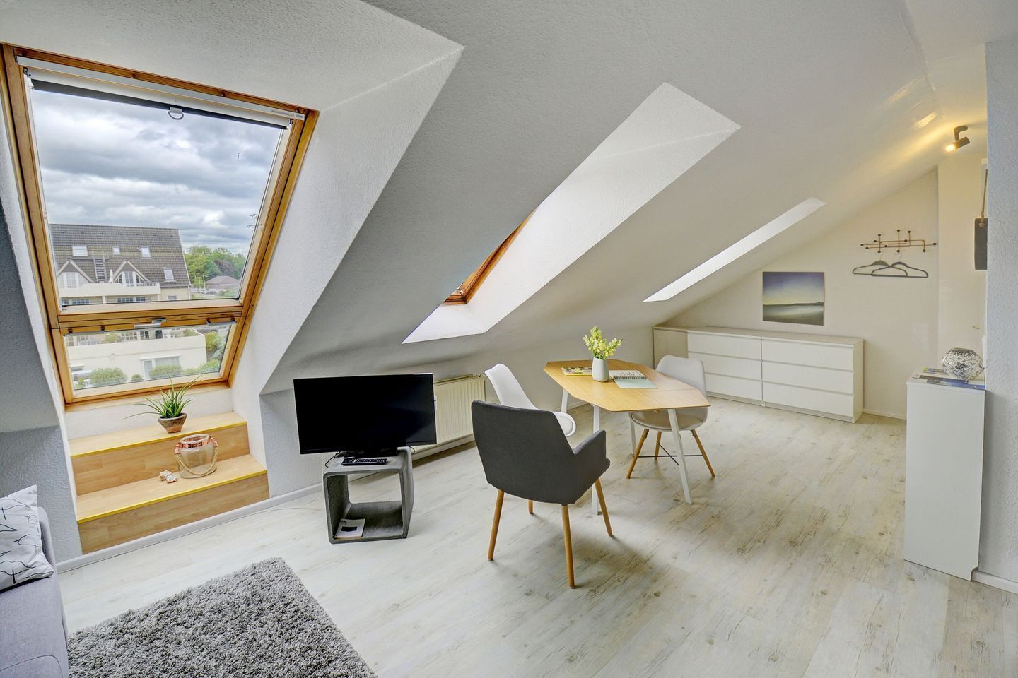 En moderne loftlejlighed med skrå vægge, et spiseområde med bord og stole, et fjernsyn og udsigt over kvarteret.