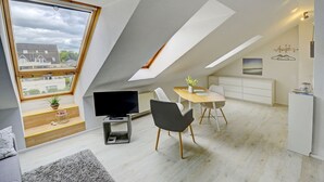 A modern loft apartment with a sloped ceiling, a dining area with a table and chairs, a TV, and a view of the neighborhood. - fewo1846 - Beach shell (App. 638) / Studio apartment with sea view on the 3rd floor - (Harrislee)