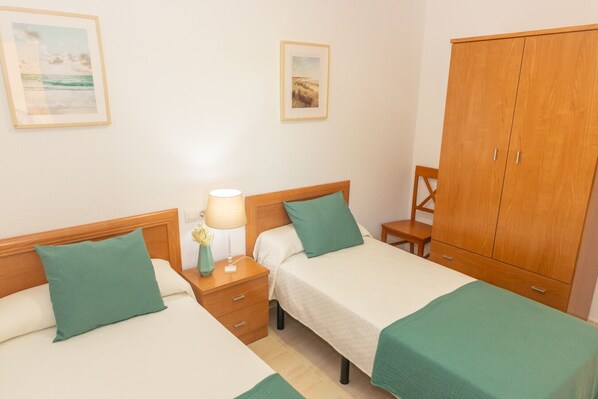 A hotel room with two beds, a wooden wardrobe, a nightstand with a lamp, and two framed pictures on the wall.
