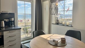 A modern kitchen with a large window overlooking a beach, a dining area with a round table, and a view of the sea. - fewo1846 - KüstenNest (App. 590) / Studio apartment with 2 balconies on the 1st floor - (Harrislee)