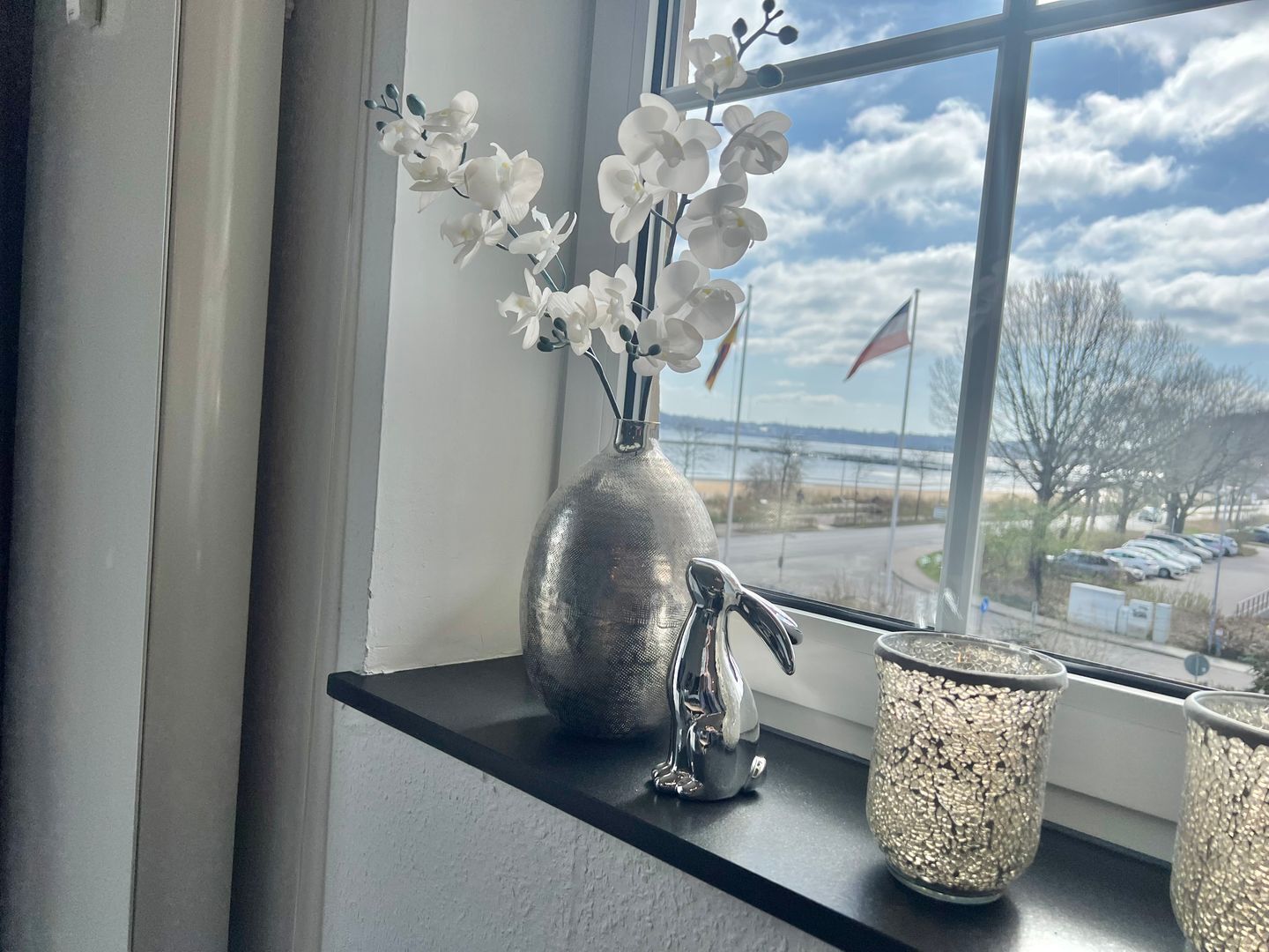 Ein Fenster mit Blick auf einen Parkplatz und eine Flagge, eine Vase mit weißen Blumen und eine dekorative Metallfigur auf einem Fensterbrett.
