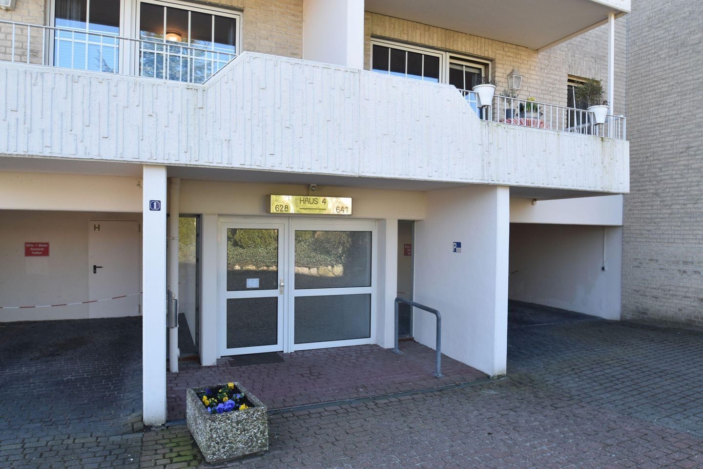 Une entrée d’immeuble avec une plaque indiquant « HAUS 4 628 641 », une entrée fermée par un portail et un petit jardinier.