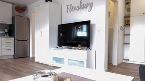 A modern kitchen with a built-in TV, white cabinetry, and a dining area with a table and decorative items. - fewo1846 - MuschelNest (App. 621) / Studio apartment with balcony on the 2nd floor - (Harrislee)