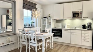 A modern kitchen with white cabinetry, a dining table with chairs, a chandelier, and a view of the outdoors through a window. - fewo1846 - MuschelNest (App. 621) / Studio apartment with balcony on the 2nd floor - (Harrislee)