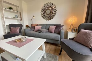 Un salon moderne avec un canapé gris, deux fauteuils, une table basse blanche et un miroir décoratif accroché au mur.