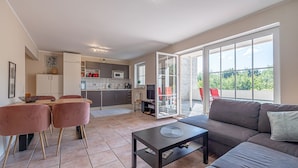 A modern living room with a sofa, a coffee table, and a dining area with chairs. There is a kitchen in the background with a sliding glass door leading to a balcony with red chairs. - fewo1846 - Fördeblick I (App. 670) / Studio apartment with balcony on the mezzanine floor (Harrislee)