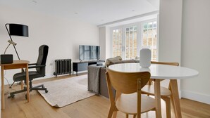 Ein modernes Wohnzimmer mit Holzboden, einem runden Esstisch, einem schwarzen Bürostuhl, einer Couch, einer Heizung und einem großen Fenster mit Blick auf eine Ziegelwand.