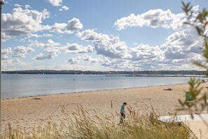 A beach with a person walking, a sandy shore, and a distant cityscape with buildings and boats on the water.