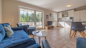 A modern living room with a blue sofa, a coffee table, and a TV. The room has large windows and a kitchen with white cabinets. - fewo1846 - Fördeblick II (Apartment 671) Studio apartment with a terrace on the mezzanine level (Harrislee)