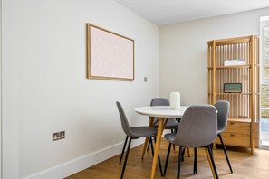 Un coin repas avec une table ronde, quatre chaises grises, un vase blanc et une bibliothèque en bois remplie de livres.