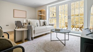A modern living room with a sofa, a coffee table, and a bookshelf. There is a large window with a view of a brick wall and outdoor seating. - Open plan Waterloo, 2BR, New build, terrace, by Blueground (London)