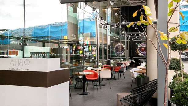 An outdoor seating area with tables and chairs, a glass wall with reflections, and a sign for ATRIO restaurant and bar. - Open plan Waterloo, 2BR, New build, terrace, by Blueground (London)