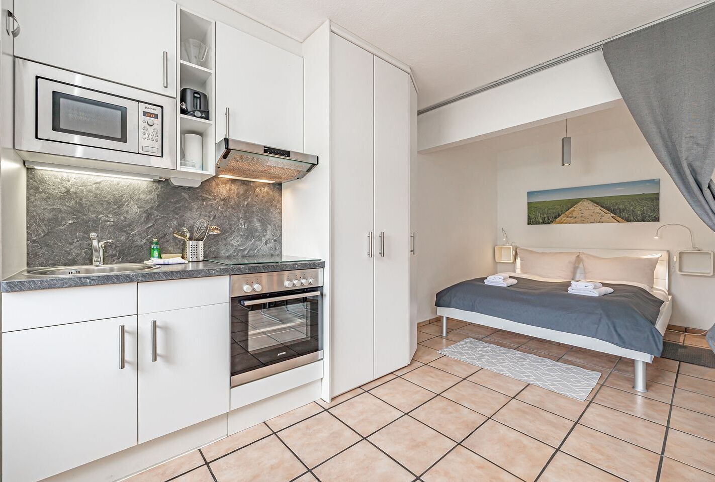 A compact hotel room with a kitchenette, a bed with a gray bedspread, a painting on the wall, and a gray curtain.