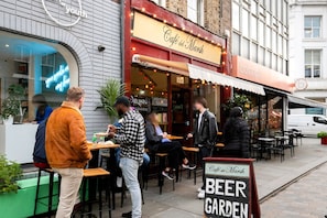 Uma área externa de estar do Café del Marsh, com mesas e cadeiras, uma placa indicando ser um jardim de cervejas, e pessoas jantando e socializando.