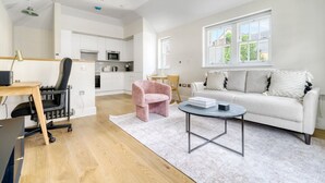 A modern living room with a white sofa, a coffee table, and a pink chair. There is a kitchen area in the background with white cabinets and a microwave. - Open plan Waterloo, 1BR, New build, terrace, by Blueground (London)