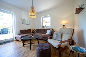 Een gezellige woonkamer met een bruine leren bank, een houten salontafel, een fauteuil met kussens en een hanglamp.