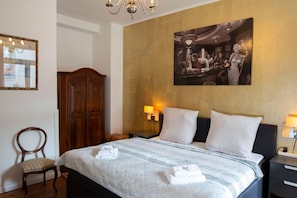 Habitación de hotel con cama, dos lámparas de noche, un armario de madera, una silla y un cuadro en la pared.