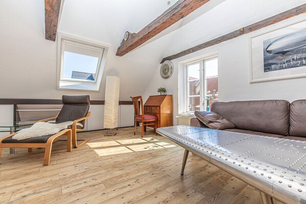 A room with a wooden floor, a brown leather sofa, a glass table with a white furry rug, a wooden rocking chair, and a metal massage table.