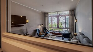 Un soggiorno moderno con televisione, due poltrone blu, un divano grigio e un'ampia finestra con vista su alberi e palazzi.