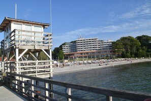 Un hotel frente al mar con una torre de salvavidas, una playa con sombrillas y un edificio grande al fondo.