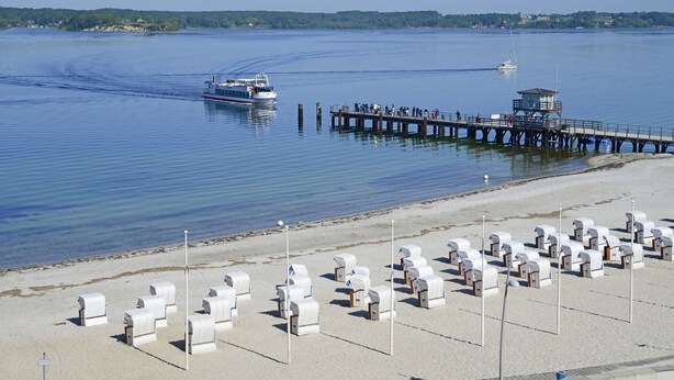 A pier extending into calm waters with a boat and a lighthouse in the distance. - fewo1846 - At the Beach - elegant studio apartment with balcony and sea view - (Glücksburg (Ostsee))