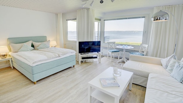 A modern hotel room with a bed, a sofa, a TV, and a dining area with a view of the sea. - fewo1846 - Fjordview (App. 18) - fewo1846 - Fjordview / modern studio apartment (Glücksburg (Ostsee))