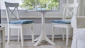 A room with two white chairs with blue cushions, a small white table, and a window with a view of trees. - fewo1846 - Fjordview (App. 18) - fewo1846 - Fjordview / modern studio apartment (Glücksburg (Ostsee))