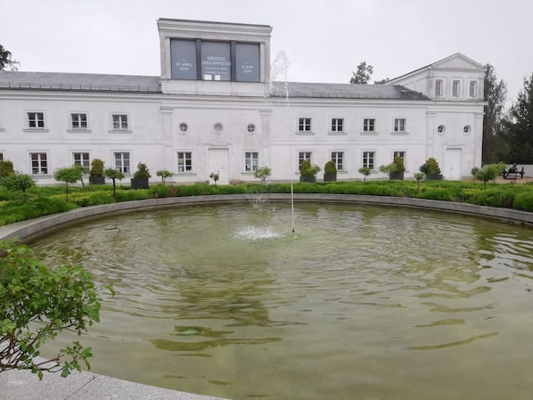 Un grand bâtiment blanc, avec une fontaine au centre d’un bassin circulaire, entouré d’arbustes et d’arbres aux formes soigneusement taillées.