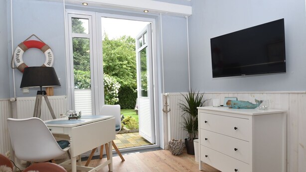 A room with a desk, chair, TV, and a view through a glass door to a garden. - fewo1846 - Pauline - fewo1846 - Pauline in Glücksburg / enchanting (Glücksburg (Ostsee))