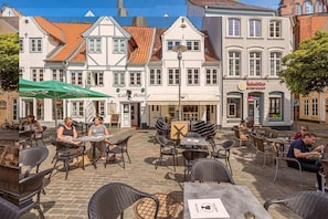 Un café en plein air avec des tables et des chaises, une enseigne indiquant « Café Alette », et un bâtiment portant une enseigne « Reischür ».