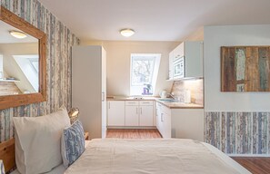 A compact bedroom with a bed, bedside table, and a mirror. Adjacent is a kitchenette with a window, sink, and cabinets.