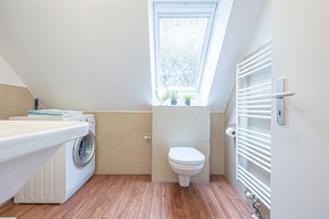 Une salle de bain moderne et compacte, équipée d’un lave-linge, de toilettes et d’une fenêtre laissant entrer la lumière naturelle.