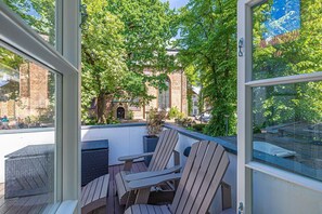 A balcony with wooden chairs and a table, overlooking a brick building and lush greenery.