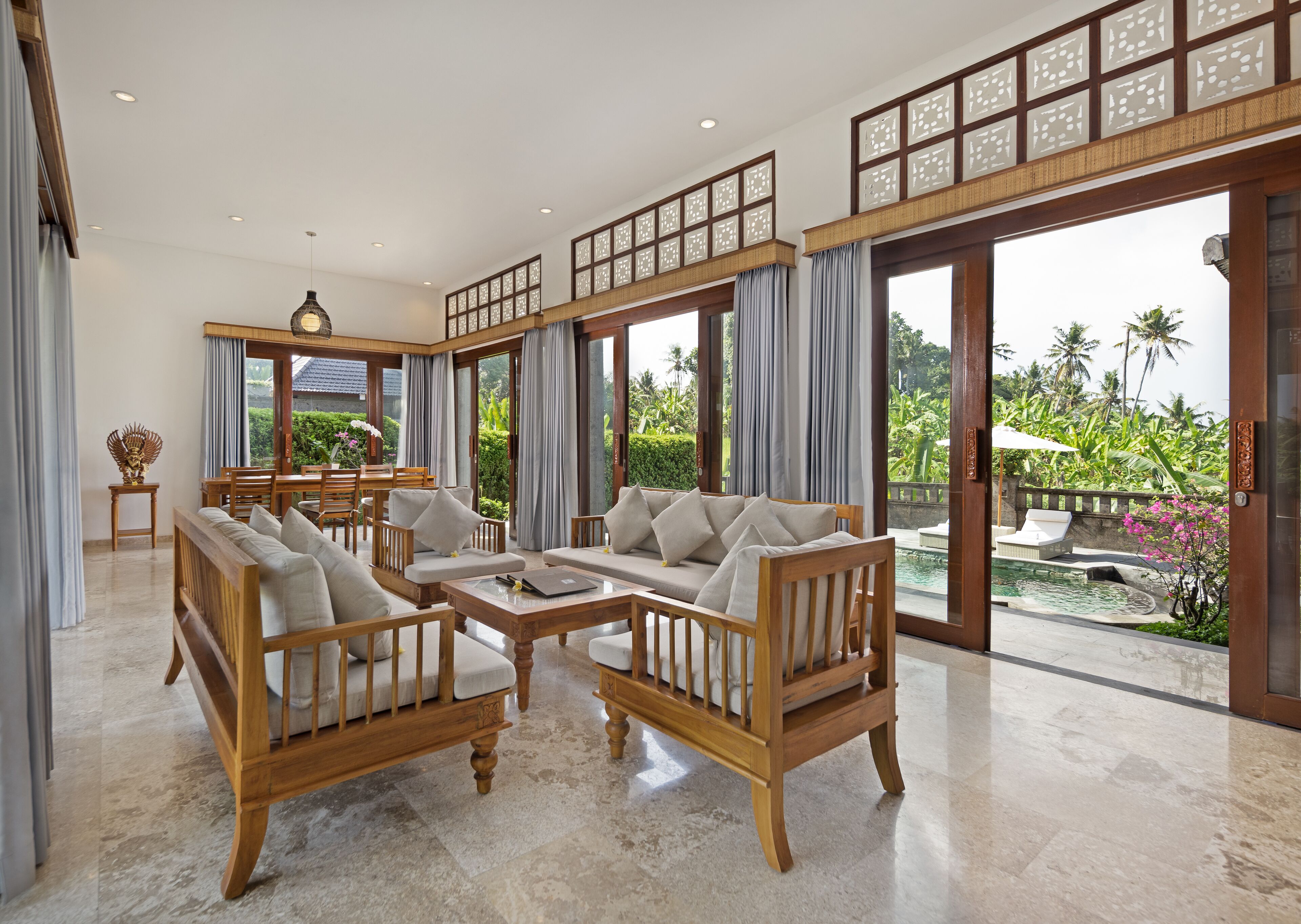 A spacious living area with wooden furniture, a dining set, and large windows offering a view of a pool and tropical garden.