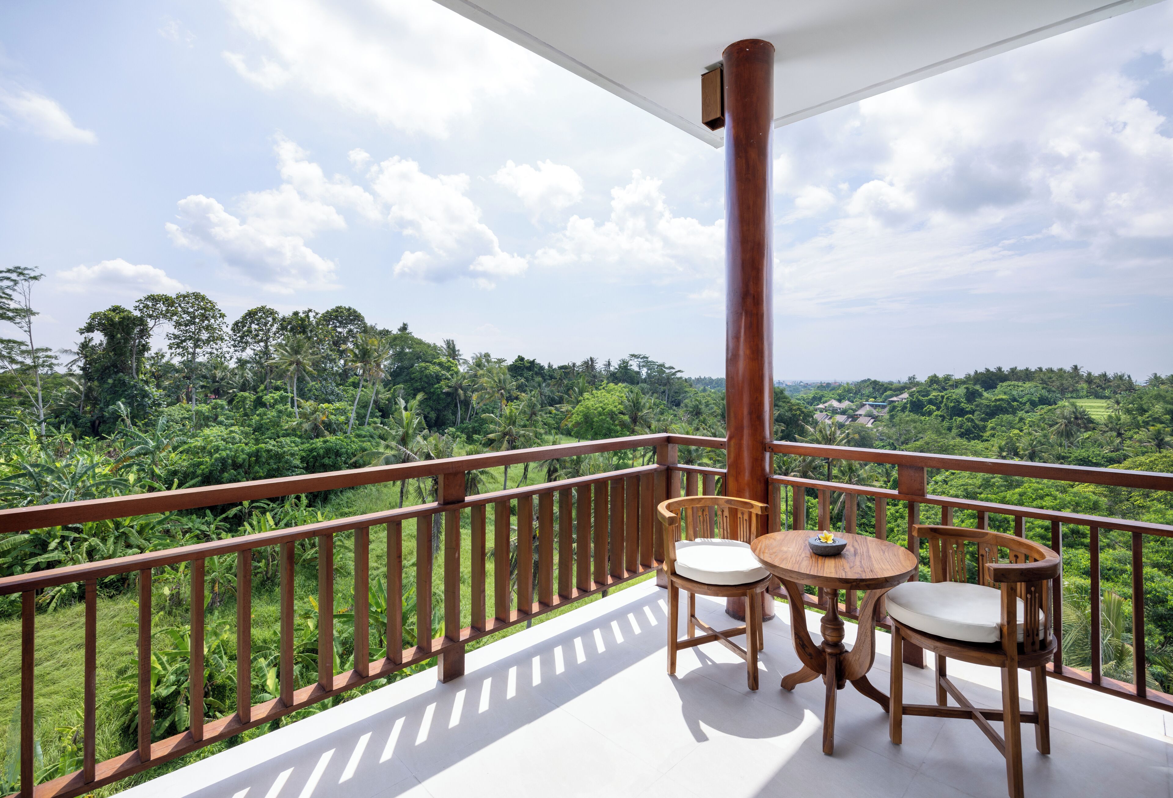 A balcony with a wooden railing, two wooden chairs, a small round table, and a view of a lush green landscape.