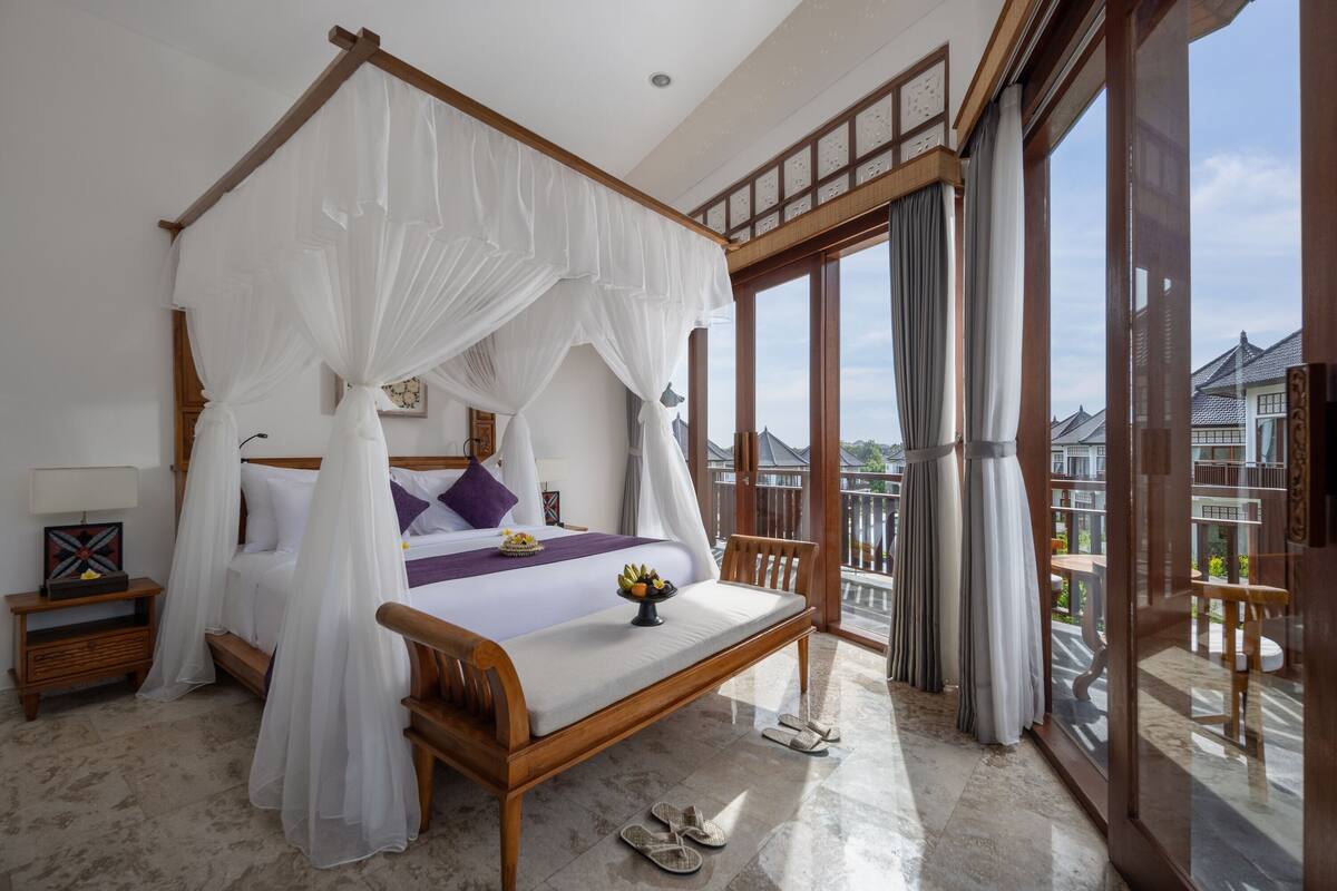 Een slaapkamer met een hemelbed, een houten bankje en een balkon met uitzicht op gebouwen.