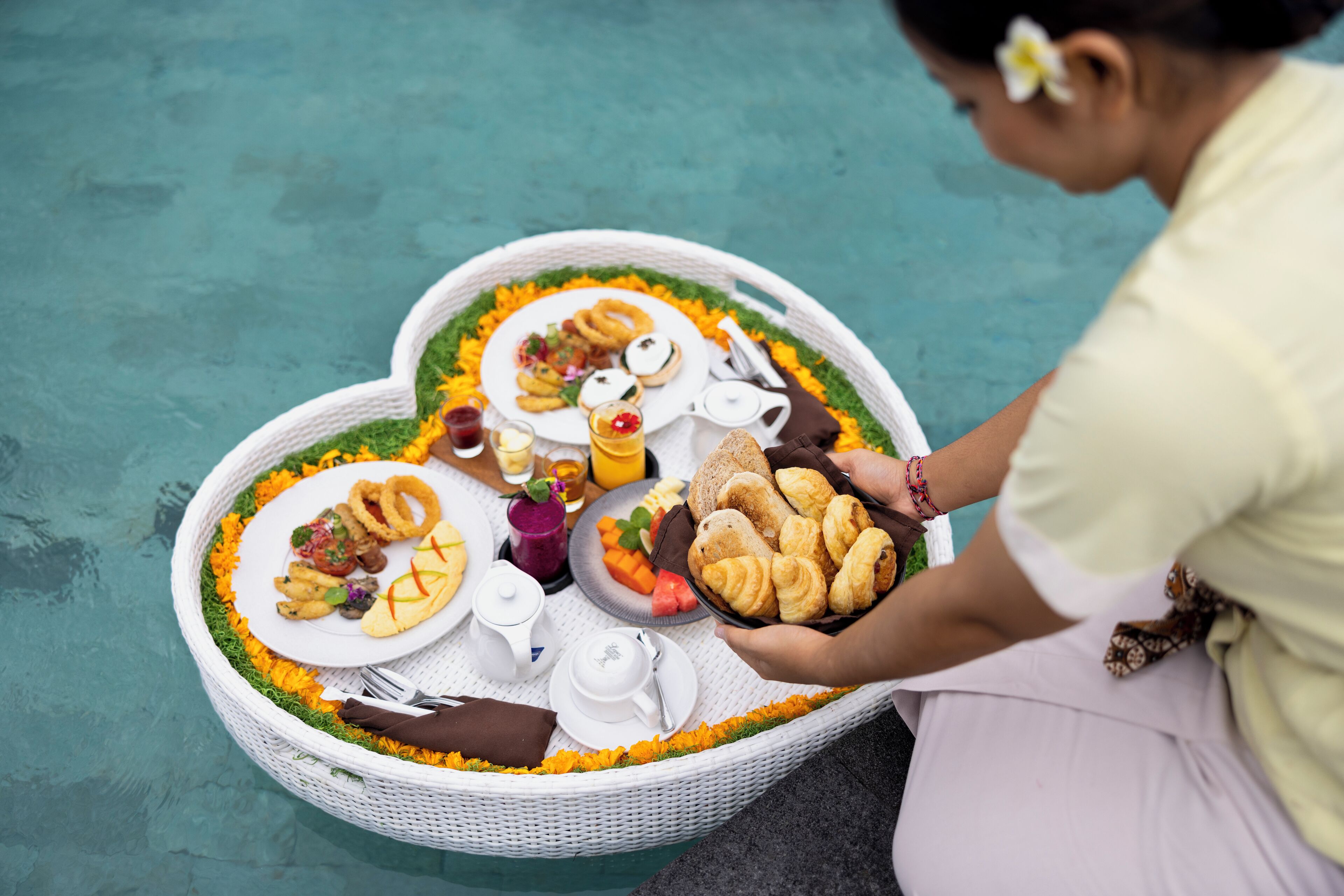 A person holding a tray with food items and a basket of pastries, with a table set with plates, cups, and a teapot, all placed on a decorative mat.
