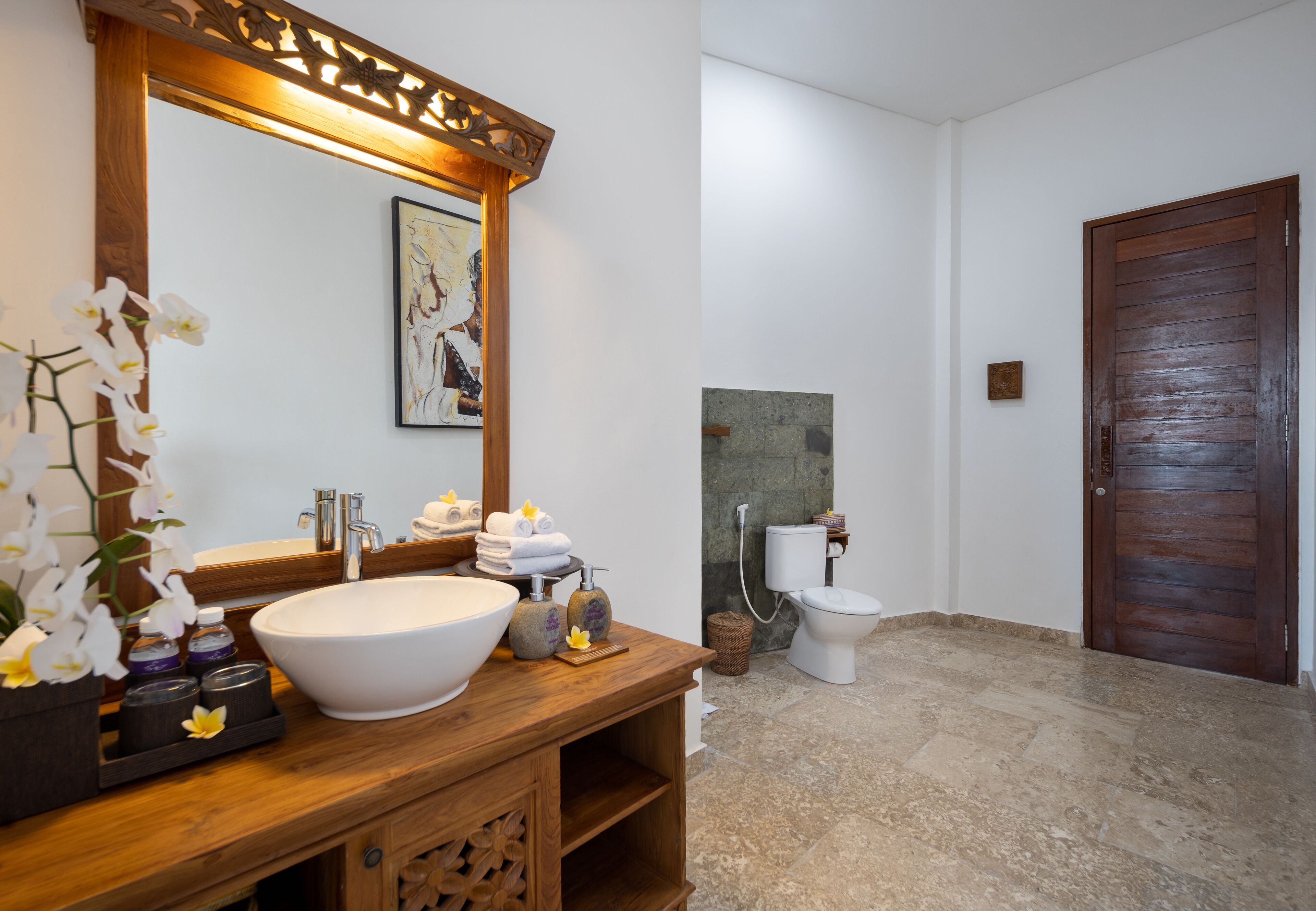 A bathroom with a wooden vanity, a large white sink, a mirror, a toilet, and a wooden door.