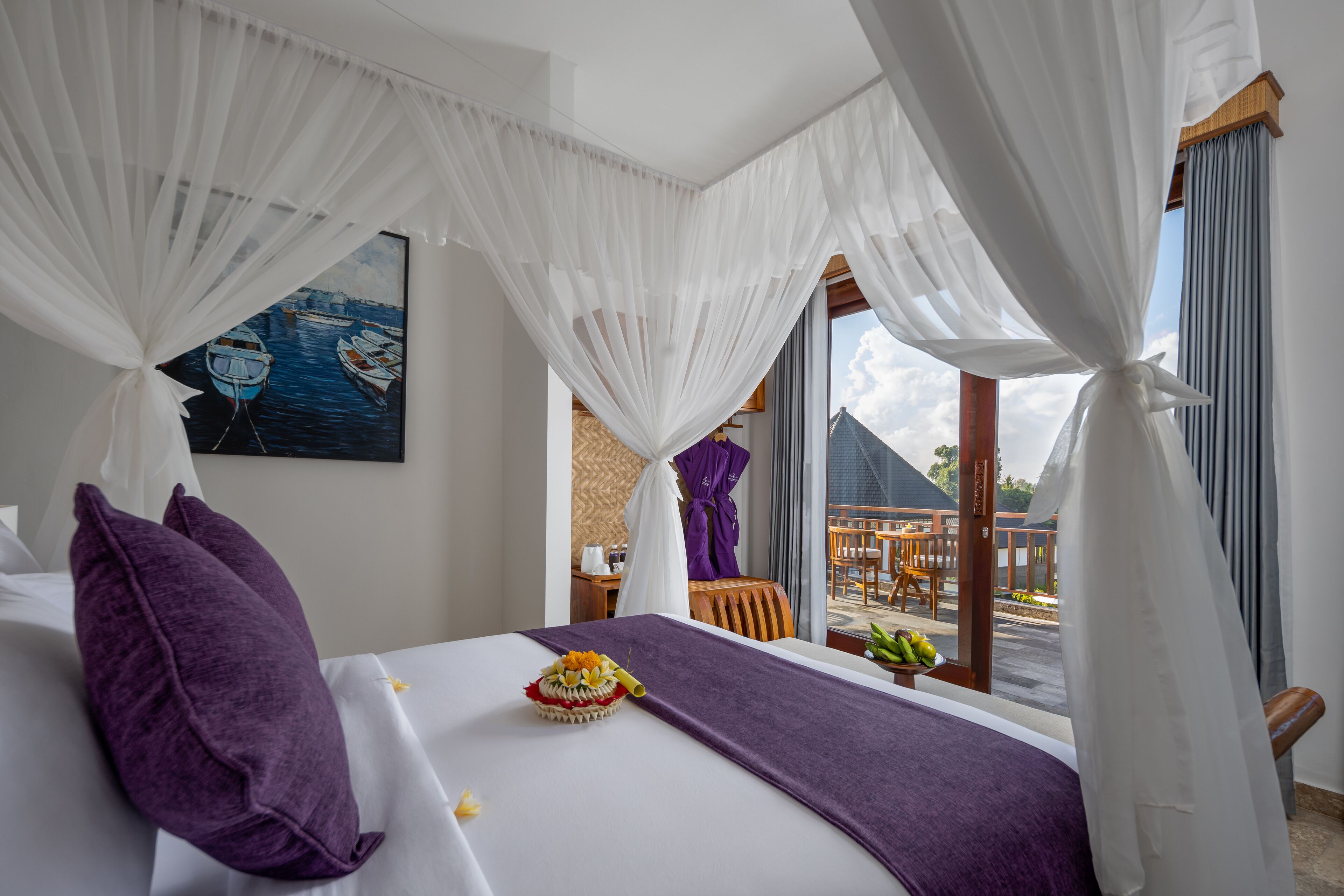 Une chambre d’hôtel avec un lit, des oreillers violets, un panier de fruits et un tableau représentant des bateaux accroché au mur.